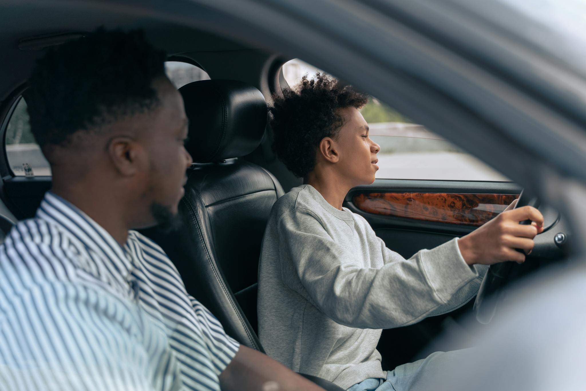 A father teaches his son to drive, sitting together in a car and discussing techniques. Side view indoors.