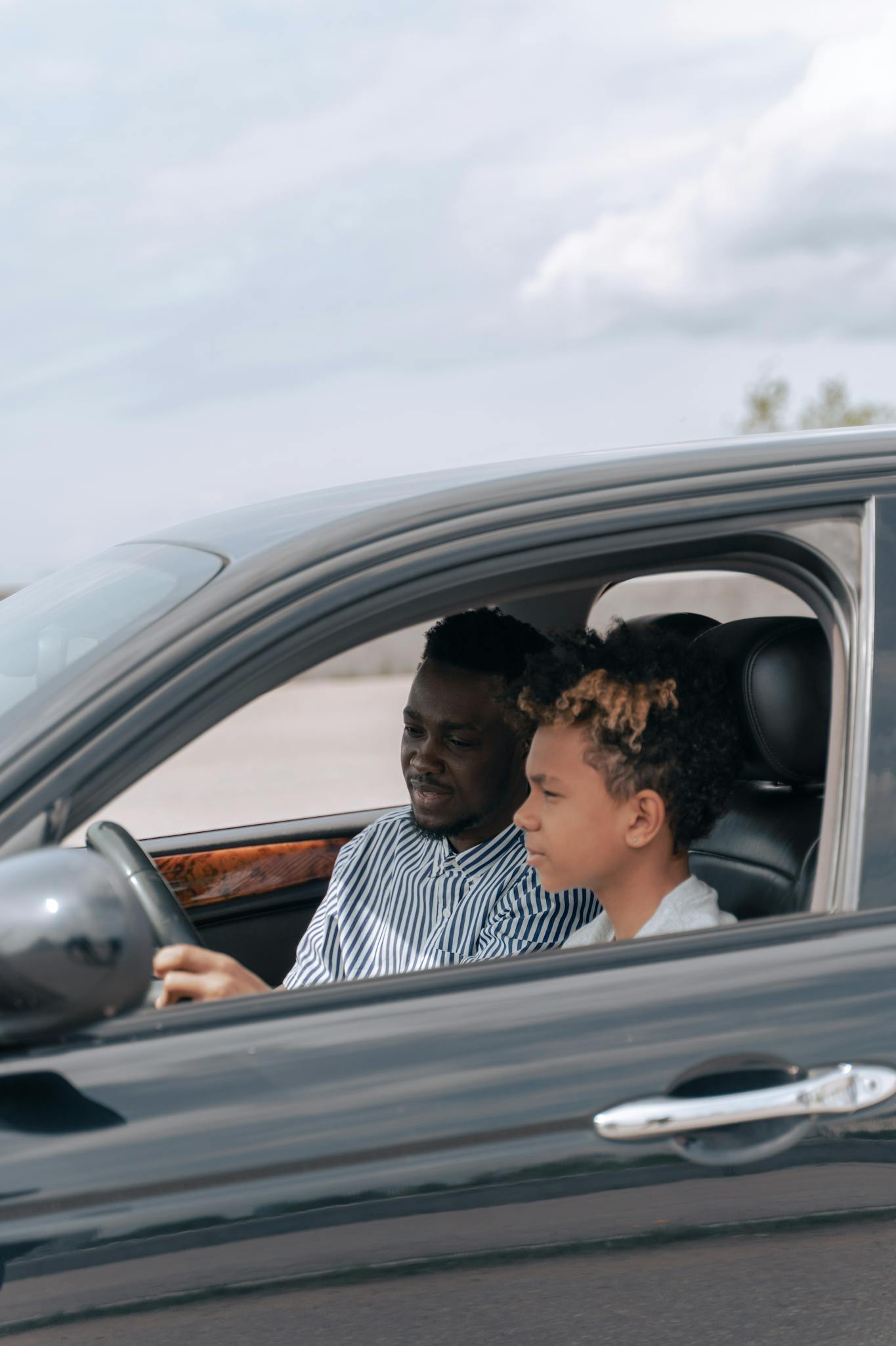 Father guides son during a driving lesson inside a black sedan on a bright day.